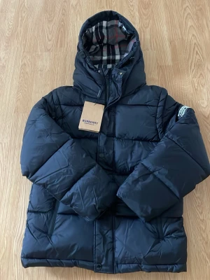  Burberry jacka - Snygg svart pufferjacka från Burberry med klassiskt rutigt foder i huvan. Jackan har en vadderad design, dragkedja och knappar framtill samt en stor huva. Perfekt för kalla dagar och riktigt stilren med märkets ikoniska detaljer.