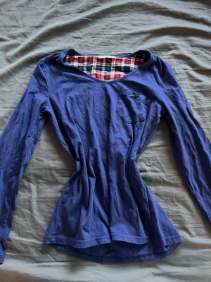Blå långärmad topp med rutigt foder - Snygg blå långärmad topp med v-ringning och rutigt foder i halsen. Liten bröstficka med broderad logga. Perfekt för dig som gillar en enkel men cool stil.