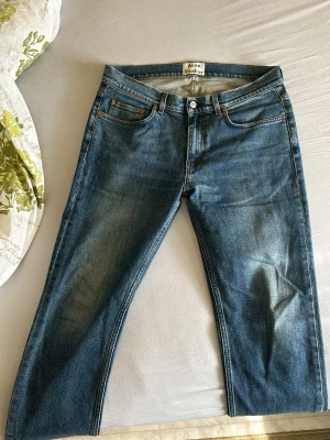 Blå raka acne studios jeans med vintagekänsla - Säljer ett par klassiska acne studios blå jeans med rak/slim passform. Jeansen har två bakfickor och är tillverkade i denim. Perfekta för en avslappnad och trendig look. Strl34/32