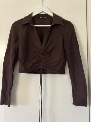 Brun croppad blus ZARA - Trendig brun croppad blus med långa ärmar och snygg dragsko-detalj framtill. Blusen har klassisk krage och är tillverkad i ett lätt, vävt material som ger en stilren look. Storlek M från ZARA 