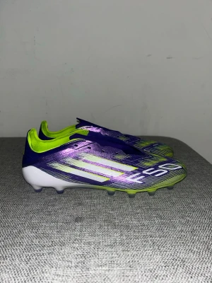 Adidas F50 lila och limegröna fotbollsskor - Säljer ett par Adidas fotbollsskor i metallic-lila med limegröna detaljer och vita ränder. Skorna har låg profil, snörningsfri design och FG-dobbar för spel på gräs. Snygg och modern look med tydlig Adidas-logga bak och på sidan.