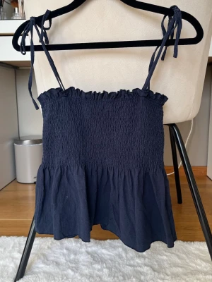 Mörkblå smockad topp med knytband - Supersöt mörkblå topp med smockad överdel och volang nedtill. Axelbanden knyts och gör att man kan justera passformen💘 Endast använd ett fåtal gånger