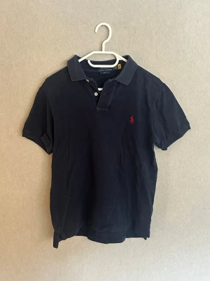 Marinblå pikétröja från Ralph Lauren - Klassisk blå pikétröja från Ralph Lauren med röd broderad logga på bröstet. Slim fit-modell med korta ärmar, krage och knappar framtill. Perfekt för en stilren och avslappnad look. Står M men sitter som Xs. Bra pris vid snabb affär😊