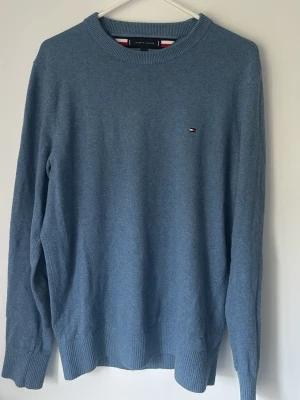 Blå stickad tröja Tommy Hilfiger - Snygg blå stickad tröja från Tommy Hilfiger med rund halsringning och diskret logga broderad på bröstet. Tröjan har långa ärmar och ribbade muddar vid ärmslut och nederkant. Perfekt för en stilren och avslappnad look.