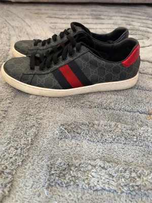 Gucci Ace Sneakers  - Snygga Gucci sneakers i svart med klassiskt GG-mönster, röda och blå ränder på sidorna samt röd och blå detalj på hälen. Skorna har svarta skosnören och vit sula. Perfekta för dig som vill sticka ut med lyxig streetstyle.