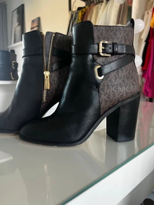 Svarta stövletter från Michael Kors - Snygga stövletter från Michael Kors i svart skinn och brunt mönstrat tyg med MK-logga. Dekorativa spännen och guldfärgad dragkedja på sidan. Klackarna är breda och stabila, perfekt för en trendig look.