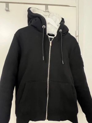 Svart hoodie från Moose Knuckles - Säljer en svart hoodie från Moose Knuckles med dragkedja, stor huva och snörning. Insidan av huvan är ljusgrå och mjuk. Hoodien har en logga i metall på ärmen och stora fickor framtill. Perfekt för dig som gillar streetwear och snygga detaljer.