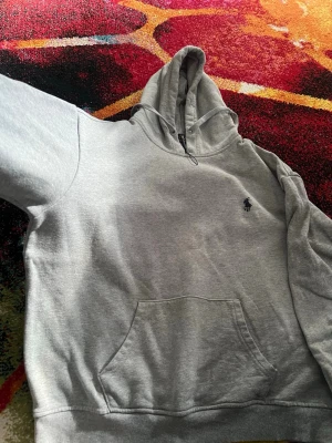 Grå hoodie från Polo Ralph Lauren - Snygg grå hoodie från Polo Ralph Lauren med klassisk broderad logga på bröstet. Hoodien har huva med dragsko och en stor magficka framtill. Perfekt för en chill och avslappnad stil. Materialet är mjukt och skönt, troligtvis bomullsblandning.