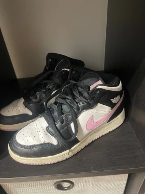 Air Jordan 1 Mid svart/vit/rosa - Snygga Air Jordan 1 Mid sneakers i svart, vitt och rosa. Skorna har klassisk design med svart läder, vita paneler och rosa swoosh. Perfekta för dig som vill sticka ut med en ikonisk och trendig look.