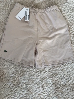 Beige shorts från Lacoste - Snygga beige shorts från Lacoste med elastisk midja och klassisk krokodillogga framtill. Perfekta för sommardagar och har en enkel, stilren design. Tillverkade i mjukt material för maximal komfort.