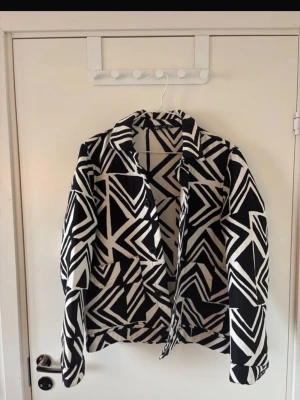 Svartvit kavaj med grafiskt mönster Zara - Snygg kavaj från Zara i storlek M med svartvitt grafiskt mönster. Rak passform, klassisk krage och långa ärmar. Perfekt för dig som vill sticka ut med en trendig och modern look. Tillverkad i ett mjukt tyg som känns bekvämt hela dagen.