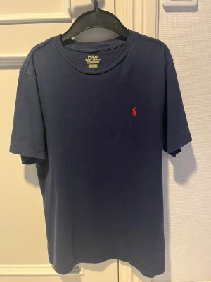 Marinblå t-shirt från Polo Ralph Lauren - Klassisk marinblå t-shirt från Polo Ralph Lauren med röd broderad logga på bröstet. T-shirten har rund halsringning och korta ärmar. Tillverkad i mjuk bomull för en skön känsla och enkel stil.