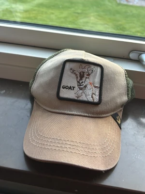 Beige och grön truckerkeps med get - Säljer en beige och grön truckerkeps från Goorin Bros med mesh baktill och böjd skärm. Framtill finns ett broderat patch med en get och texten 'GOAT'. Perfekt för dig som vill ha en unik och trendig accessoar.