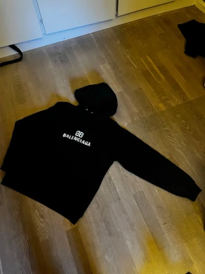 Svart Balenciaga hoodie med tryck - Svart hoodie från Balenciaga med vit pixelinspirerad logga och BB-tryck på bröstet. Klassisk modell med huva och dragsko. Perfekt för dig som gillar streetwear och vill sticka ut med ett exklusivt plagg. ÄKTA!!!