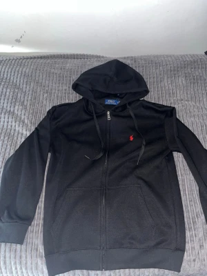 Svart hoodie från Polo Ralph Lauren - Svart hoodie med dragkedja från Polo Ralph Lauren. Klassisk design med huva och dragsnören, två fickor framtill och röd broderad logga på bröstet. Mjuk bomullskänsla och ribbade muddar vid ärmslut och nederkant.