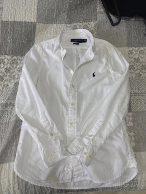 Vit skjorta från Ralph Lauren - Klassisk vit skjorta från Ralph Lauren med den ikoniska broderade loggan på bröstet. Skjortan har lång ärm, knappar framtill och vid manschetterna samt en tidlös passform. Tillverkad i mjuk bomull som känns skön mot huden.