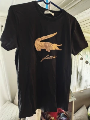 Svart Lacoste t-shirt med broderad logga/S - Svart t-shirt från Lacoste med stor broderad guldfärgad krokodil och Lacoste-signatur på bröstet. Klassisk rund hals och korta ärmar. Perfekt för dig som gillar stilrena plagg med en twist. Kan inte garantera äkthet 