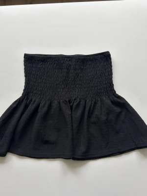 Svart kortkjol från Gina Tricot - Supersöt svart kortkjol från Gina Tricot med smockad midja och lätt utställd form. Den går att använda som topp eller kjol, vilket är prima! Storlek XS 