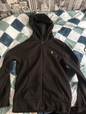 Svart hoodie från Polo Ralph Lauren - Svart hoodie med dragkedja från Polo Ralph Lauren. Klassisk design med huva, två fickor framtill och den ikoniska vita logotypen broderad på bröstet. Tillverkad i mjukt bomullsmaterial som är skönt att ha på sig. Perfekt för en avslappnad och stilren look.
