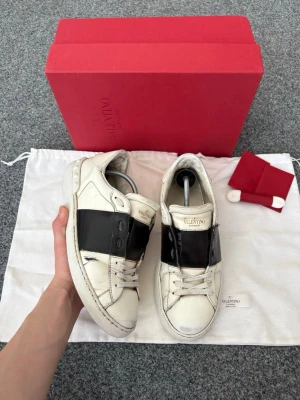 Valentino opens - Snygga vita sneakers från Valentino med bred svart läderdetalj över mitten. Klassisk låg modell med rund tå, vita skosnören och diskret logga på plösen. Skorna är i läder och har en chunky sula som ger en cool vibe.