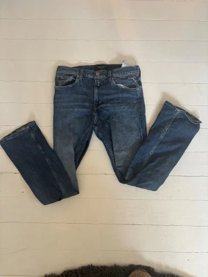 Blå raka jeans från Replay - Säljer ett par klassiska blå jeans från Replay med rak passform och fem fickor. Jeansen har snygga slitningar och en läderpatch med Replay-logga bak i midjan. Tillverkade i denim med en autentisk tvättad look. Passar längd 158- 170 cm