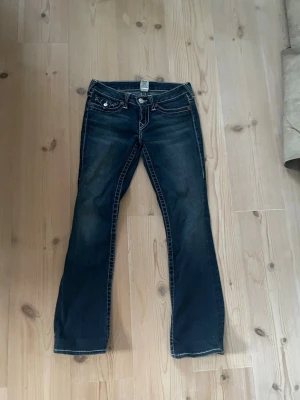 Mörkblå True Religion jeans - Säljer ett par mörkblå jeans från True Religion med kontrastsömmar och ikoniska fickdetaljer bak. Utsvängda/ lite raka ben och snygg tvätt med slitningar. 