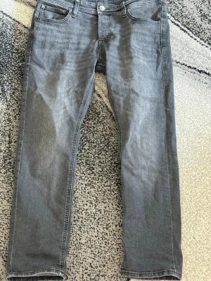 Grå jeans från Jack & Jones - Snygga grå jeans från Jack & Jones med klassisk femficksdesign och raka ben. Jeansen har en diskret tvättad look och är tillverkade i ett mjukt denimtyg som ger en skön passform. Perfekta för en avslappnad stil.
