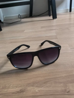 Svarta solglasögon med mörk blått och svart glas (Grish Glasögon) - Snygga svarta solglasögon med klassisk, fyrkantig form och mörka, tonade glas. Perfekta för att ge en cool vibe till din outfit. Bågarna är i plast och modellen är tidlös och passar de flesta stilar.