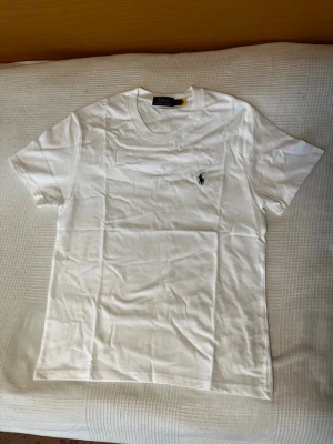 Vit t-shirt från Polo Ralph Lauren - Klassisk vit t-shirt från Polo Ralph Lauren med rund halsringning och korta ärmar. Ikoniska broderade loggan i svart på bröstet. Tillverkad i mjuk bomull som känns skön mot huden. Perfekt basplagg för en clean och stilren look. Storlek medium