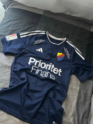 Blå Djurgården matchtröja Adidas - Snygg mörkblå matchtröja från Djurgården med vita detaljer och klubbmärke på bröstet. Tröjan har tryck från sponsorer som Prioritet Finans och Nordic Wellness samt klassiska Adidas-ränder på axlarna. Perfekt för fotbollsträning eller att bära på matchdag.