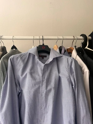 Blå randig skjorta från Dressman  - Snygg blå skjorta med diskreta ränder från Dressman. Klassisk krage och långärmad modell, perfekt för en stilren look. Skjortan har knappar framtill och är tillverkad i ett lätt och bekvämt material.