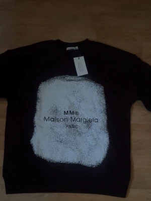 Maison Margiela tröja  - Cool svart sweatshirt från Maison Margiela med stor vit grafisk print och logga på bröstet. Tröjan har rund halsringning och är tillverkad i mjukt material, perfekt för en trendig och avslappnad stil.