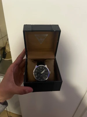 Svart klocka från Guess i originalbox - Stilren klocka från Guess med svart urtavla och silverfärgade detaljer. Klockan har ett klassiskt runt boett och svart armband. Levereras i original Guess-box vilket gör den extra lyxig. Perfekt accessoar för att lyfta din outfit.