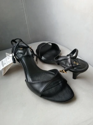 Nya Zara  sandaler med taxklack strl 41 - Stilrena svarta sandaler från Zara med smala remmar och låg klack. Skorna har fyrkantig tå och justerbar ankelrem med guldfärgat spänne. Tillverkade i slätt konstläder som ger en trendig och elegant look. 100% läder
