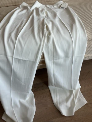 Vita vida kostymbyxor i storlek 40 - Snygga vita kostymbyxor med vida ben och pressveck. Byxorna har hög midja och stängs med dragkedja fram. Perfekta för en clean och stilren look. Materialet är mjukt och följsamt, vilket ger en skön känsla hela dagen.