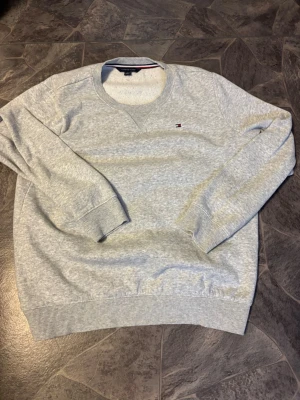 Grå sweatshirt från Tommy Hilfiger - Snygg grå sweatshirt från Tommy Hilfiger med rund halsringning och diskret broderad logga på bröstet. Tröjan har ribbade muddar vid ärmslut och nederkant samt klassisk passform. Perfekt för en avslappnad och stilren look.