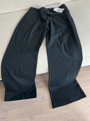 Vida svarta kostymbyxor med knytband - Snygga svarta kostymbyxor med vida ben och knytband i midjan. Byxorna har hög midja och pressveck framtill för en clean look. Perfekta för dig som gillar en avslappnad men ändå dressad stil.