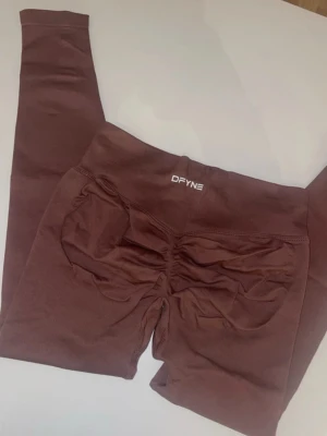 Bruna scrunch-leggings från DFYNE - Snygga bruna leggings från DFYNE med scrunch-detalj bak som framhäver formen. De är sömlösa och har hög midja för en bekväm passform. Perfekta för gymmet eller chill. Materialet är stretchigt och mjukt, vilket gör dem riktigt sköna att bära.