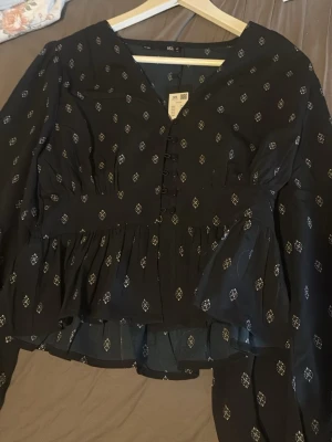 Svart blus med mönster från 157 - Svart blus från 157 med små vita mönster, knappar framtill och volang nertill. Blusen har lång ärm och lätt v-ringad hals. Tunt och luftigt material som ger en skön känsla. Perfekt för dig som gillar en trendig och avslappnad stil.