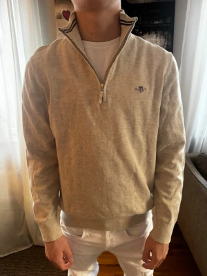 Beige halvzip tröja från GANT - Säljer en beige halvzip tröja från GANT i Small med diskret logga på bröstet. Tröjan har ribbade muddar och krage med svarta ränder, tillverkad i mjuk bomull. Perfekt för dig som gillar klassisk och stilren design.