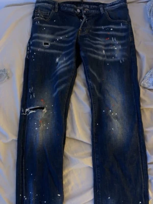 Dsquared2 jeans - Säljer ett par mörkblå dsquared2 jeans med coola slitningar och färgstänk i vitt och rött. Jeansen har klassisk femficksdesign och raka ben. Perfekt för dig som gillar en edgy och kreativ look. Materialet är denim.