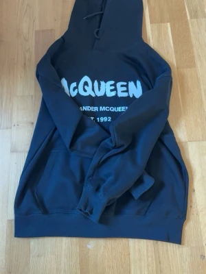 Svart hoodie från Alexander McQueen - Cool svart hoodie från Alexander McQueen med stor vit logotyp på bröstet och text under. Klassisk huva med snörning och stor magficka framtill. Perfekt för dig som vill ha en streetstyle-look med premiumkänsla.