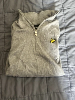 Grå halvzip tröja från Lyle & Scott - Snygg grå långärmad tröja från Lyle & Scott med halvzip och klassisk gul fågel-logga på bröstet. Tröjan har ribbade muddar och är tillverkad i mjukt material som passar perfekt till en avslappnad stil.     Pris kan diskuteras 