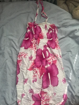 Vit och rosa blommig sommarklänning - Säljer en vit klänning med stora rosa blommor och smock upptill. Klänningen har tunna axelband och är perfekt för varma dagar. Den är lätt och luftig med ett tropiskt mönster som verkligen sticker ut.