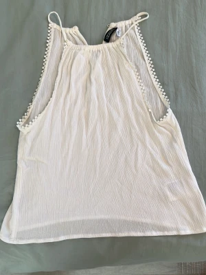 Vit axelbandslös linnetopp Gina Tricot - Säljer en vit ärmlös topp från DIVIDED HM med tunna axelband och pärldetaljer i tyg längs sidorna. Toppen har en lätt rynkad struktur. Perfekt för varma dagar och enkel att matcha med jeans eller kjol.