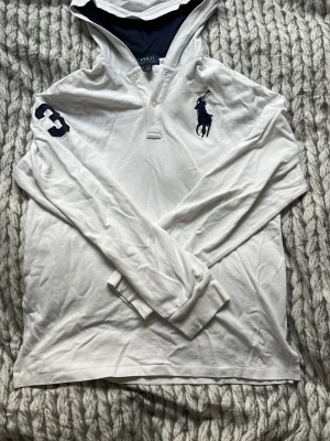 Vit långärmad polotopp från Polo Ralph Lauren - Vit långärmad polotopp med huva från Polo Ralph Lauren. Stort mörkblått logotryck på bröstet och siffran 3 på ärmen. Klassisk krage med knappar och mjukt bomullsmaterial. Perfekt för dig som gillar sportig och stilren look. Storlek XL i barnstorlek men passar som S 