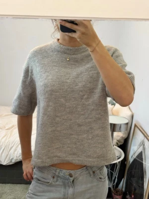 Grå stickad tröja Gina Tricot - Mjuk och stilren grå stickad tröja från Gina Tricot med korta ärmar och rund halsringning. Tröjan har en rak passform och ribbade kanter vid ärmslut och nederkant. Perfekt att matcha med jeans för en avslappnad look.