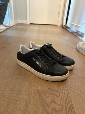 Svarta Plein Sport sneakers med silverdetalj - Snygga svarta sneakers från Plein Sport med vit sula och silverdetalj på hälen. Skorna har klassisk låg profil, svarta skosnören och logotyp på sidan. Materialet är läder och syntet, perfekt för dig som vill ha en stilren look. OBS! PRIS KAN DISKUTERAS 