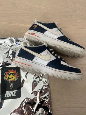 Nike Air Force 1 Low blå/vit Special edition - Säljer ett par Nike Air Force 1 Low sneakers i blått och vitt med klassisk Swoosh-logga på sidan och coolt solmotiv på hälen. Skorna har vita snören, orange detaljer och är tillverkade i läder och syntetmaterial. Perfekt för dig som vill ha en unik och stilren look. Nike loggan ändras också färg i solen från vit till gul. Pris kan diskuteras 