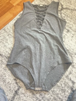 Grå body med snörning framtill - Snygg grå body med djup V-ringning och dekorativ snörning framtill. Tillverkad i mjukt stretchigt tyg som sitter skönt mot kroppen. Perfekt att styla med jeans eller kjol för en trendig look.